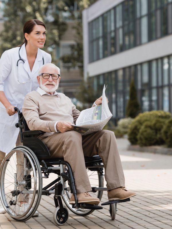 geriatric nurse walking with elderly disabled man in wheelchair with newspaper outdoors