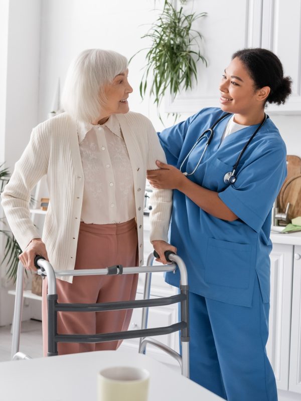 happy multiracial nurse in blue uniform supporting senior woman with grey hair walking with help of