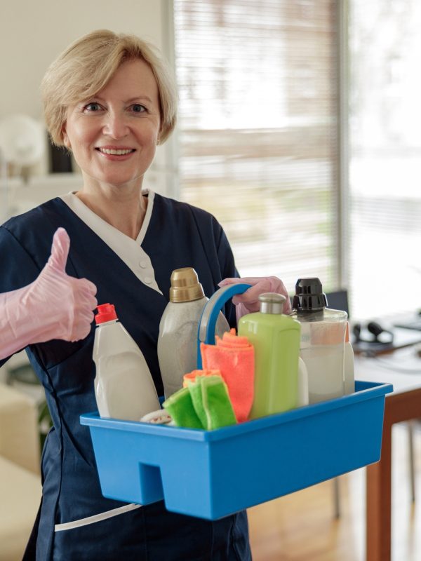 Housemaid in uniform for cleaning apartment holds box with detergents and rags and looking camera